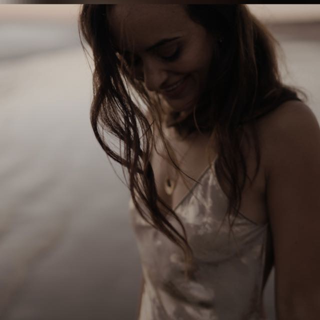 Retrato en tonos cálidos de María Garrido Moreno sonriendo mientras camina junto al mar al atardecer, con un vestido satinado claro y ambiente íntimo y cinematográfico.